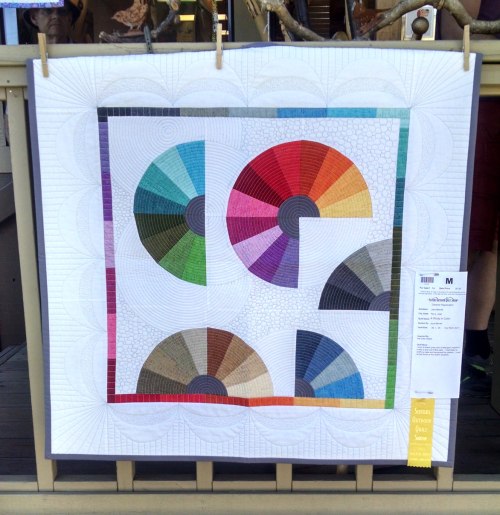 Hanging quilt with white background. The quilt depicts segments of color wheels in many shades.