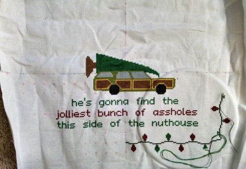A photograph of a partially completed cross stitch project. There is a car with a christmas tree on top, and the words "he's gonna find the jolliest bunch of assholes this side of the nuthouse" underneath.
