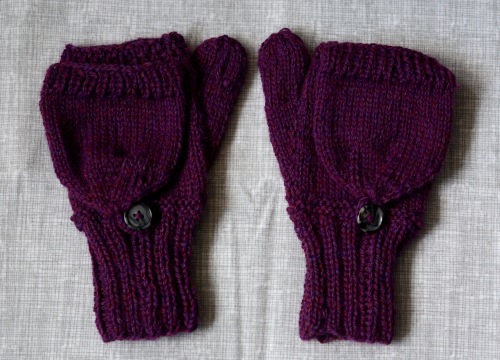 A pair of knitted mittens side by side, shown from the back. The tops fold down to become fingerless mittens, and are secured with a button